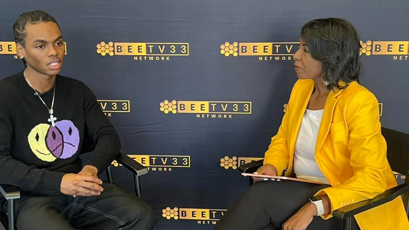 Black man wearing a cross necklace and hair braids talks with a Black woman in a yellow blazer in front of a BeeTV banner background.