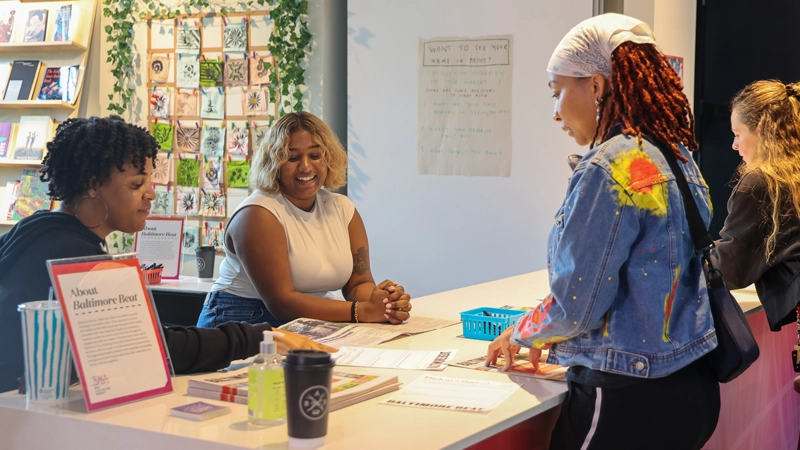 Two Black women behind a table at a community event, speaking with another Black woman getting information about Baltimore Beat.