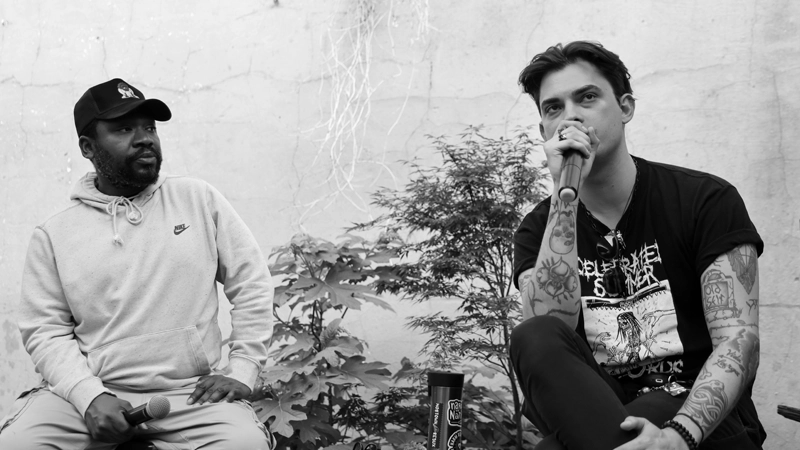 Black man in baseball cap and hoodie sweats listens as white man with tattoos speaks in a microphone.