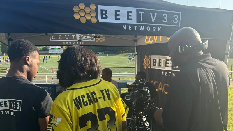 Back view of two Black men and one Black woman wearing a sports jersey in front of a BeeTV tent. They're looking into cameras together.