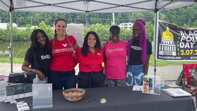 Five women smiling side by side under a tent at a community tabling event. A Black Policy Day banner is in the background.
