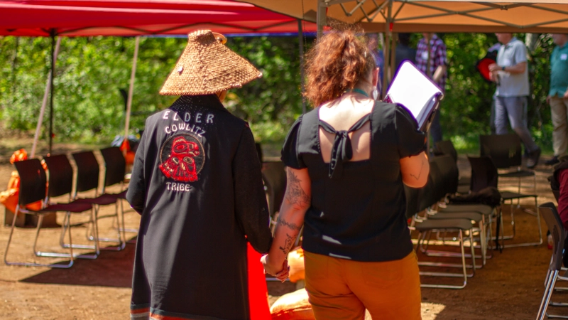 Back view of red haired woman walking and holding hands with Native woman wearing a straw hat and jacket embroidered with Elder of Cowlitz Tribe.