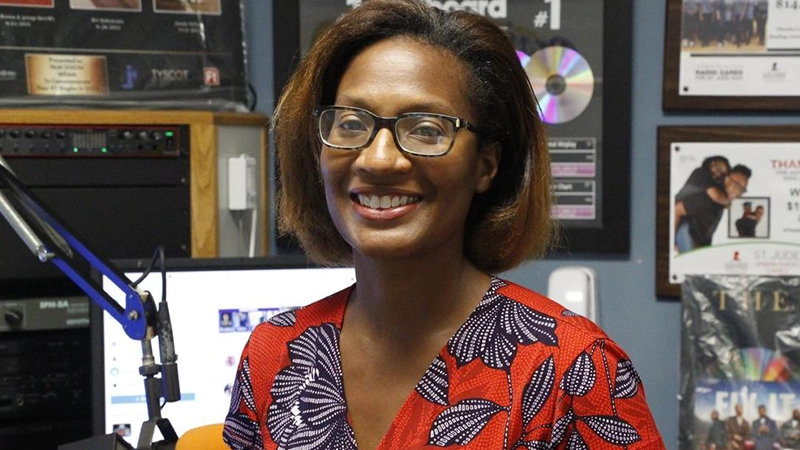 Smiling Black woman wearing a vibrantly printed dress in a working news studio.
