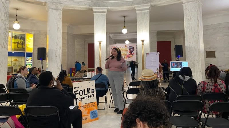 Woman speaks into a microphone standing in a marble hall room while seated people listen.