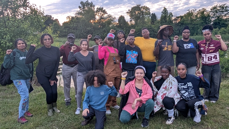 Group of Black and Brown people with strong, resolute expressions gathered in front of a pond outdoors, holding up their fists in solidarity.