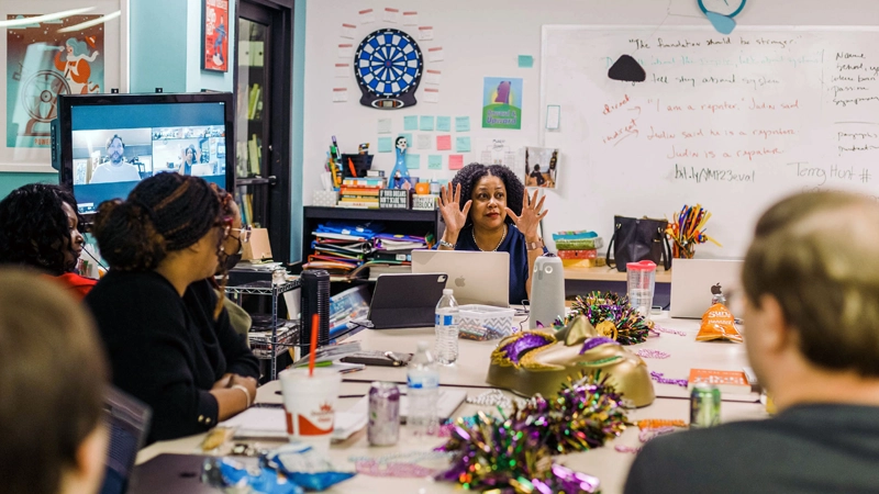 Diverse staff members discuss news around a conference table in a lively newsroom office