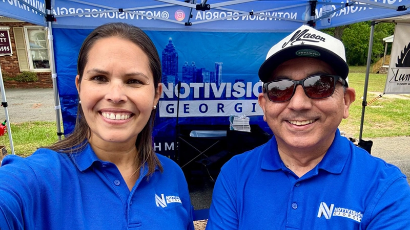 Woman and man wearing blue Notivision polo shirts at a community event in front of a Notivision banner tent.