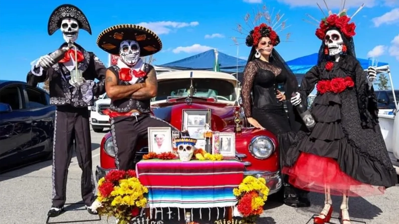 Adults dressed up in colorful traditional Day of the Dead costumes in front of a red classic car.
