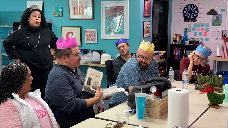 Staff wearing paper crowns in a lively newsroom office. A man is showing his coworkers a book and smiling.