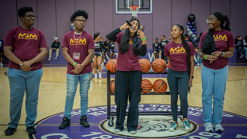 High school students wearing MIM t-shirts standing on a basketball court speaking into a microphone