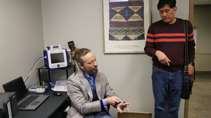 Two men in an office setting, one man is seated and demonstrating a tool on his finger while the other man looks on.