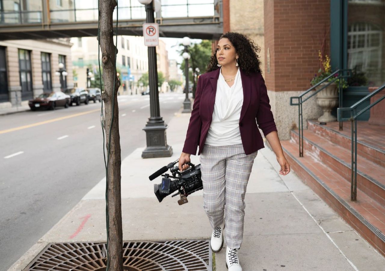Black woman with long hair walking down an urban street with a video camera in her hand