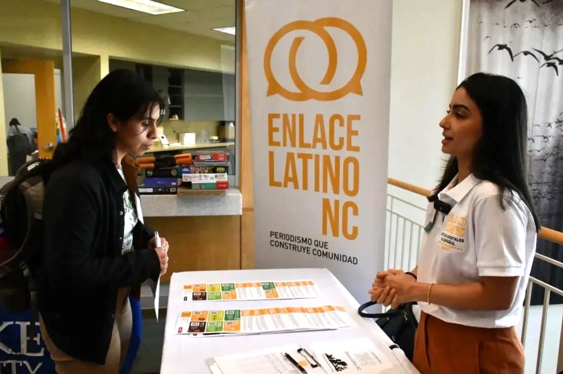 Women speaking at a table event with Enlace Latino NC banner in the background