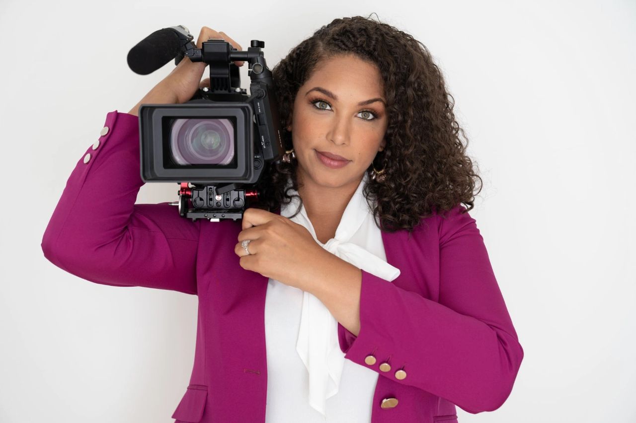 Black woman with long hair holding a video camera on her shoulder.
