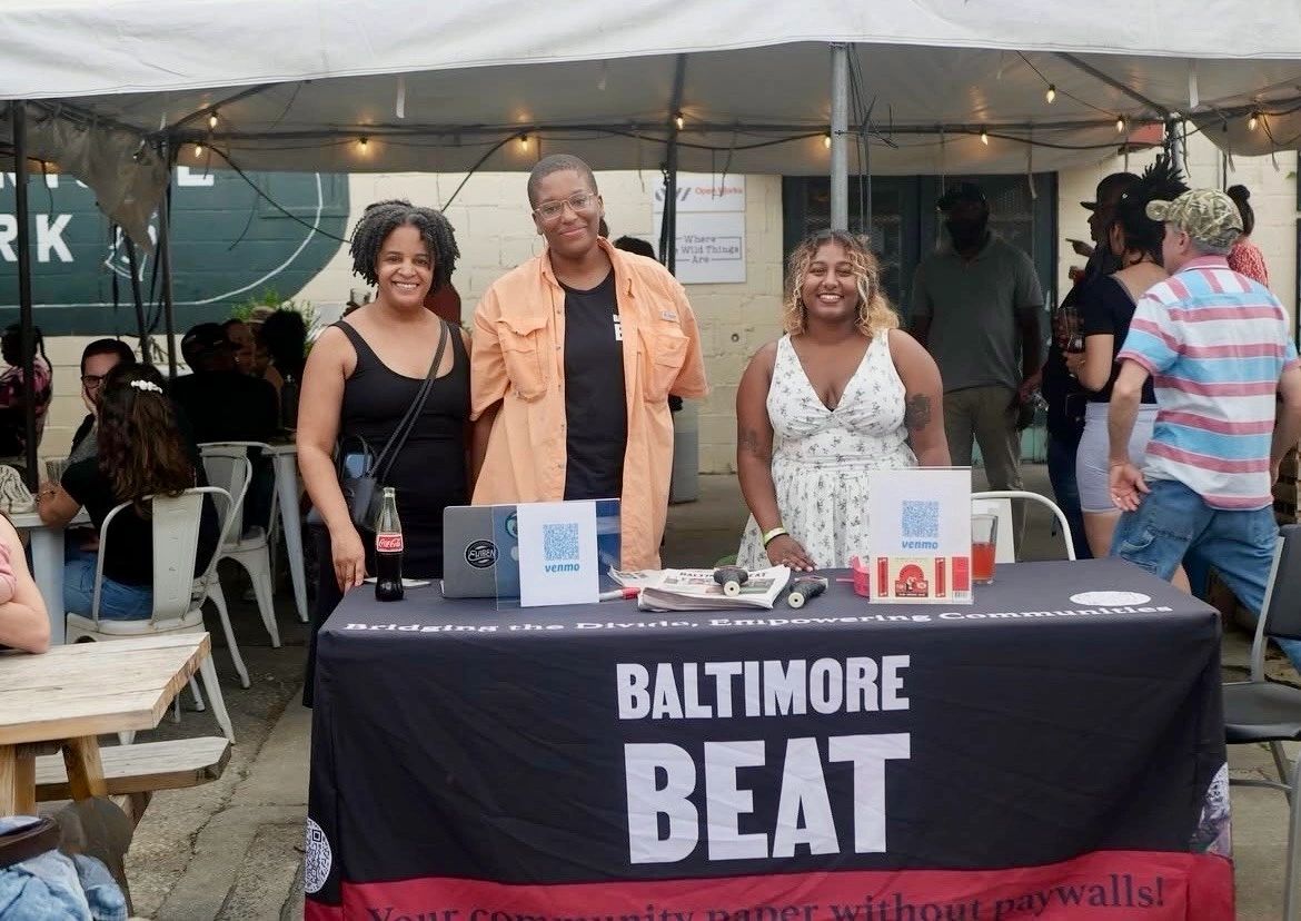 Smiling staff members behind a tablecloth that says Baltimore Beat
