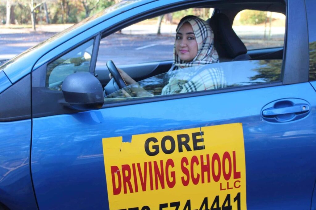 Afghan woman with head scarf behind the wheel of a car with a Gore Driving School sign on the door