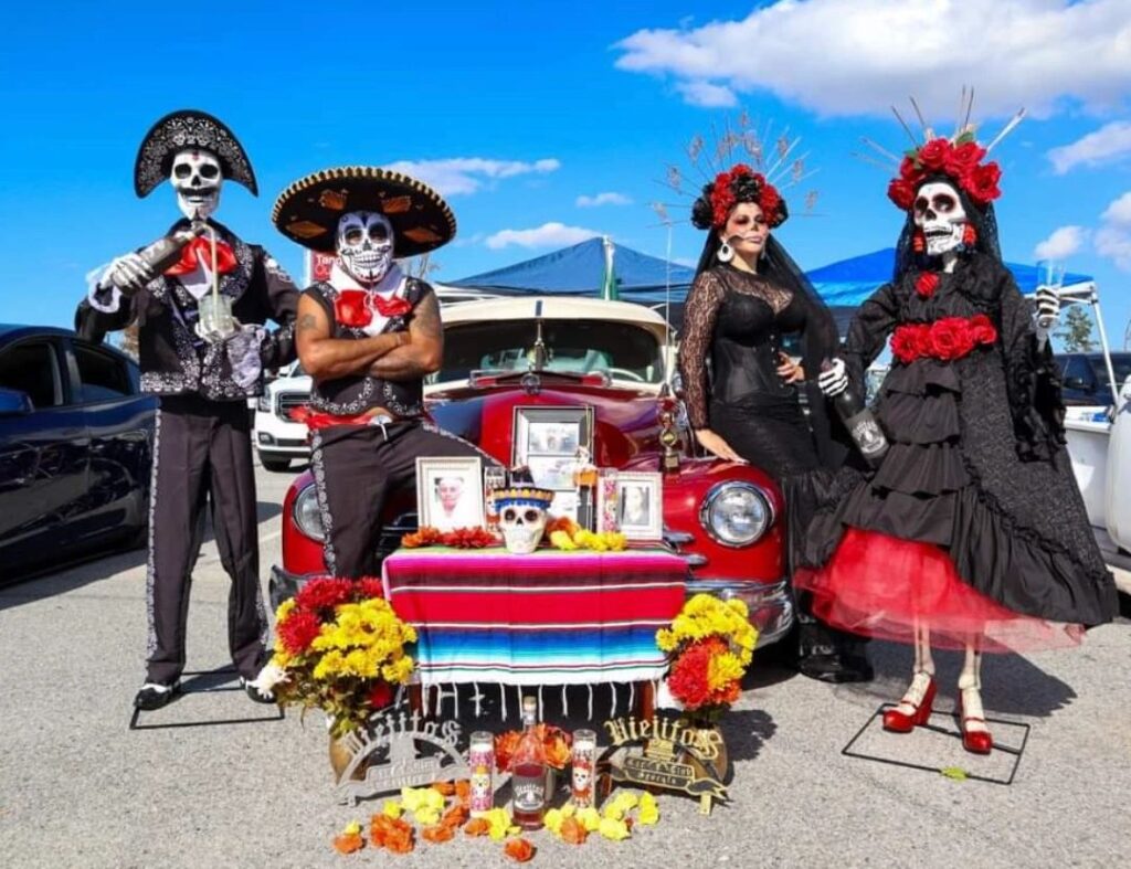 In 2023, Pasa La Voz hosted a Día de los Muertos Festival at Tanger Outlets in Pooler, GA. People standing in front of an old-fashioned car wearing colorful Day of the Dead costumes