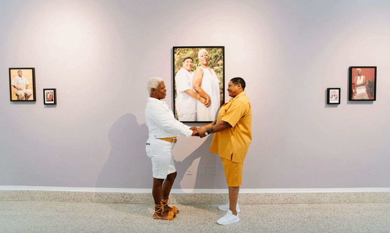 Two black women in a gallery holding hands and looking at each other fondly.