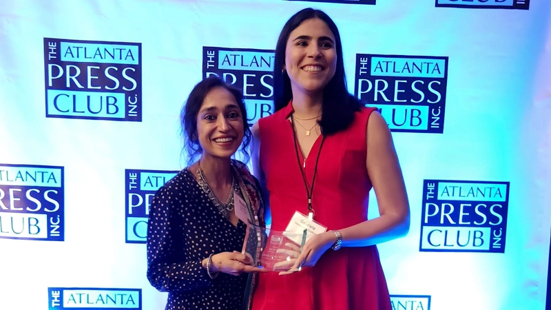 Two women smiling and holding an award in front of a Atlanta Press Club banner.