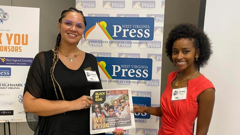 Two Black women holding a WV Pride newspaper in front of a West Virginia Press banner.