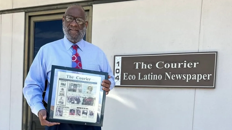 Mature Black man holding a framed copy of The Courier Eco Latino newspaper in front of a Courier Eco Latino sign outside their office.