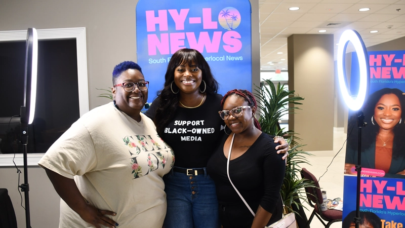Three Black women smiling in front of a Hy-Lo News banner and ring lights.