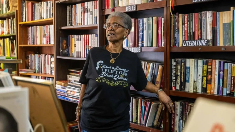 Mature Black woman in a Black is Beautiful tshirt standing in front of a bookshelf full of books.