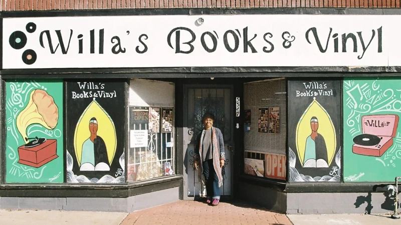 Mature Black woman in a knit cap, sweater and scarf smiles standing in front of a bookshop with funky illustrations in the window and "Willa's Books & Vinyl" painted at the top.