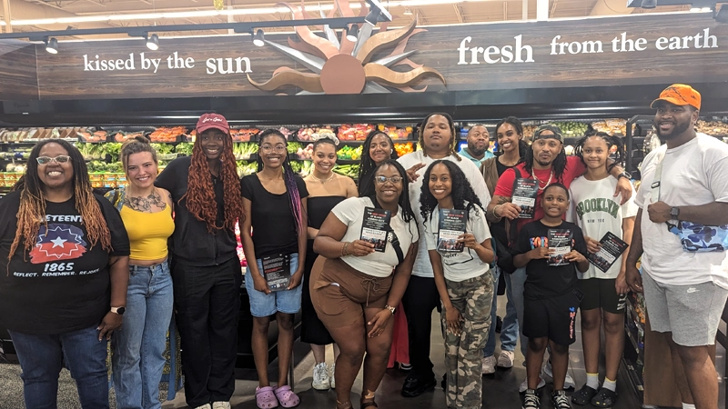 Group of mostly Black people of different ages and genders smiling in the produce department of a grocery store.