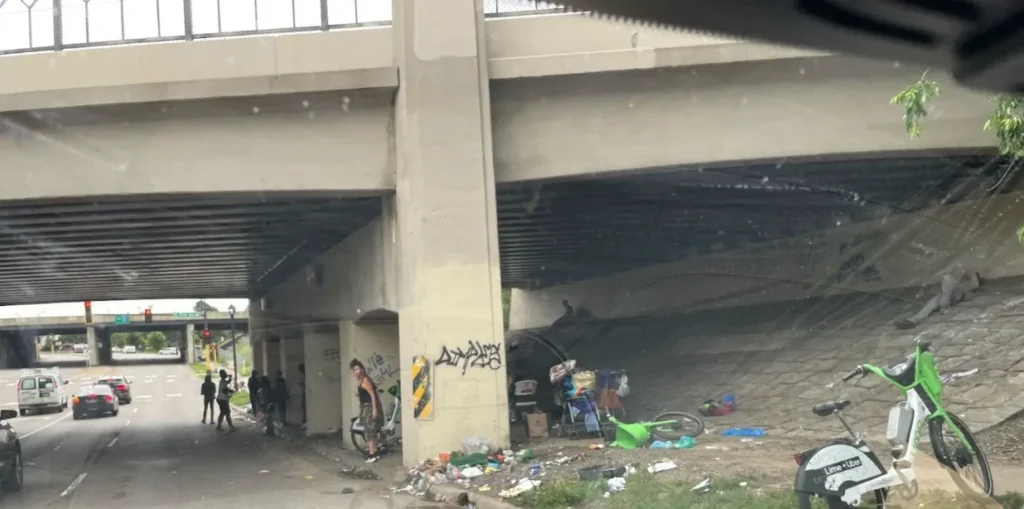 People, bikes, camping supplies and trash under a bridge overpass next to a highway.