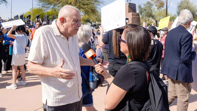 Female reporter at an outdoor community event, holding a microphone to record a mature man speaking.