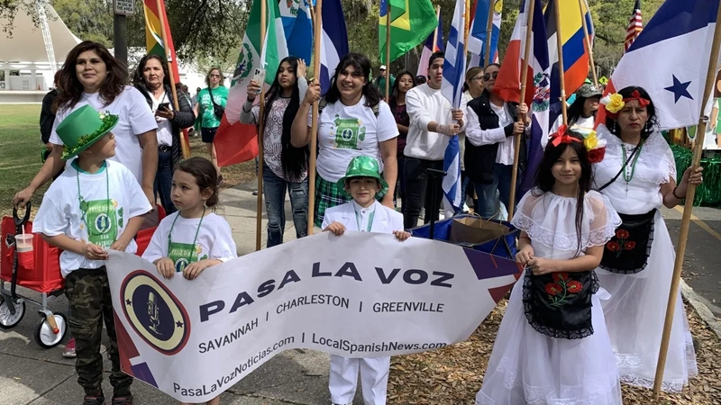 Children and adults wearing white walk in a parade carrying flags from different central and south american countries.
