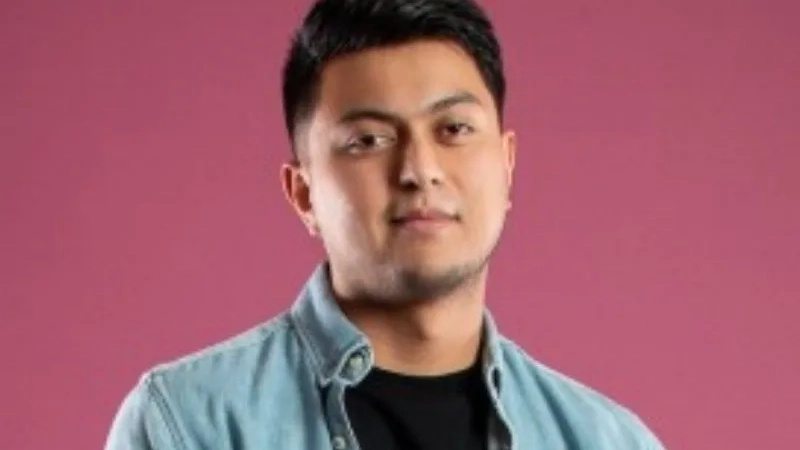 Young Latino man with a half smile wearing a denim shirt.