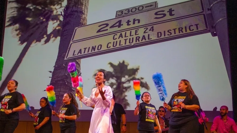 Dancers sing and clap in front of a street sign that says 24th Street Calle 24 Latino Cultural District