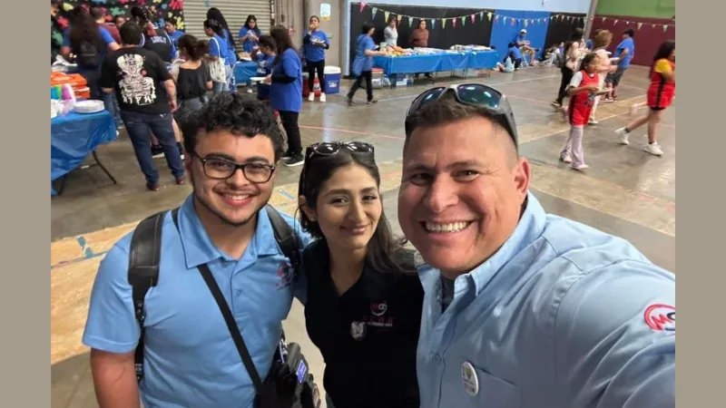 Two men and a woman take a smiling selfie in a busy gymnasium at an event
