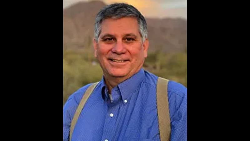 Mature light skinned man wearing a blue collared shirt and tan suspenders smiling with a mountain landscape in the background.