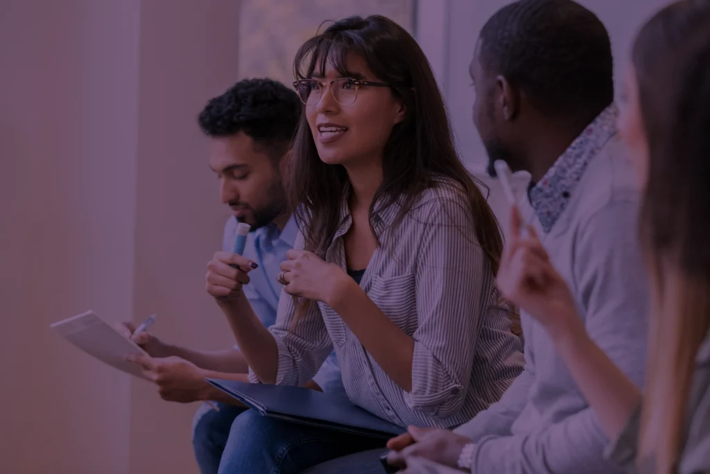 Latina reporter sitting next to other reporters from different ethnic backgrounds asking a question to someone off camera.