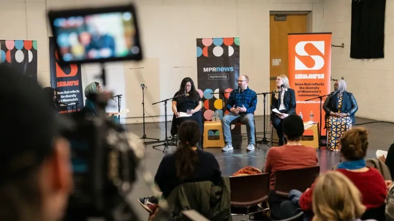 Five people on a stage speaking in front of banners and presentation screens with audience in the foreground.
