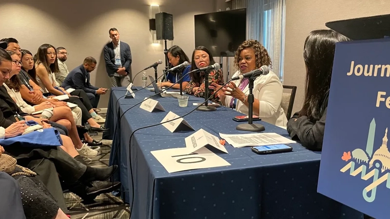 Four women of color at a table with microphones at the front of the room, with one Black woman talking. Attendees listen with interest.