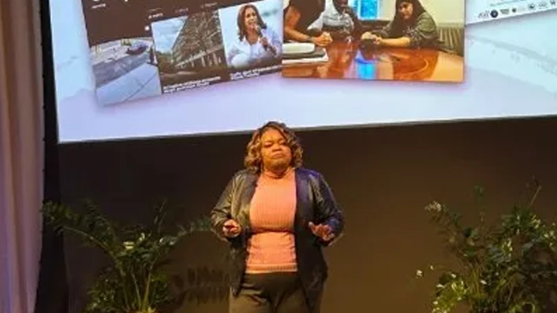 Black woman in leather jacket speaking to a large audience in front of a presentation screen