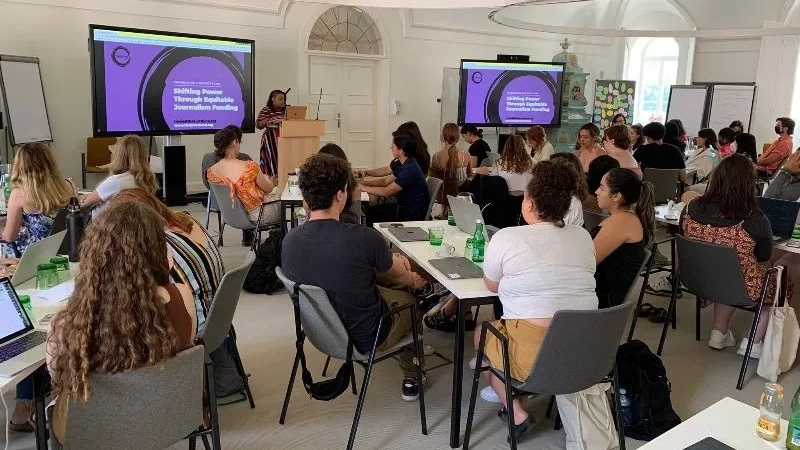 Black woman giving a presentation in front of presentation slides to people seated around tables.