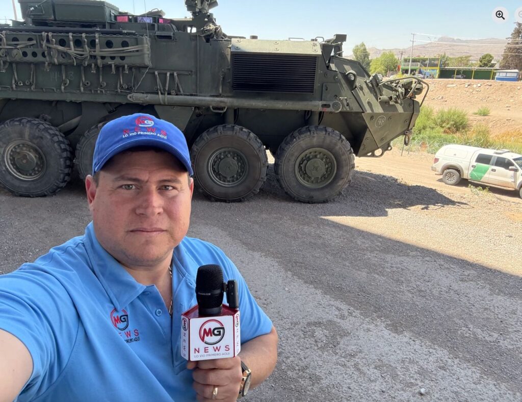 Man with blue baseball cap and a serious expression holds a microphone while standing in front of a tank.
