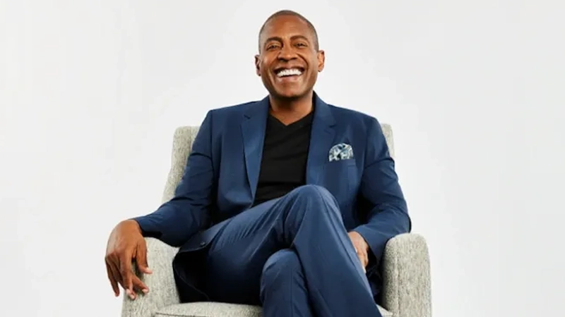 Black man in a blue suit sitting on a chair with legs crossed smiling